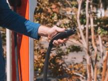 Hand holding an electric vehicle charger at a station outdoors with autumn trees. (Foto: Kindel Media / Pexels)