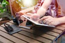 Person using a tablet outdoors alongside a camera, phone, sunglasses, and notebook on a wooden table. (Foto: Porapak Apichodilok / Pexels)