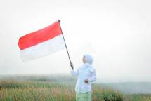 Smiling woman in hijab holding Indonesian flag in misty outdoor field. (Foto: Ruly Nurul Ihsan / Pexels)