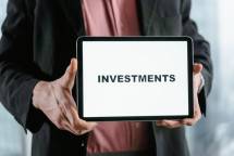 A businessman in a suit holds a tablet displaying the text 'INVESTMENTS'. (Foto: Tima Miroshnichenko / Pexels)